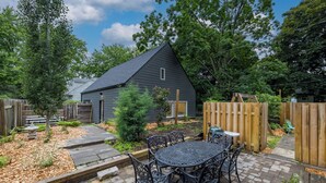 Ein Garten mit einem dunklen Haus, einem Holzzaun, einem Metalltisch mit Stühlen und einem kleinen Gartenbereich.