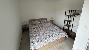 A bedroom with a bed, a bedside table with a lamp, a wall-mounted shelf with clothes, and a power outlet. - Balcon sur la Rivière Face à la Vieille Ville (Périgueux)