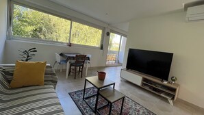 A modern living room with a sofa, a TV, a dining table with chairs, and a balcony with a view of trees. - Balcon sur la Rivière Face à la Vieille Ville (Périgueux)