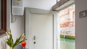 Ein Zimmer mit einem kleinen Tisch, einer Topfpflanze und einem Glas roter Flüssigkeit. Eine weiße Tür führt zu Blick auf einen Kanal.