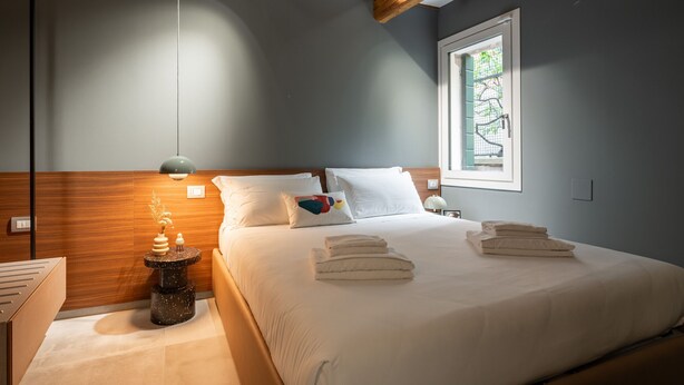 Ein ordentlich bezogenes Bett mit gefalteten Handtüchern, ein Holz-Kopfteil, ein Nachttisch mit Lampe und ein Fenster mit Blick auf Grün.