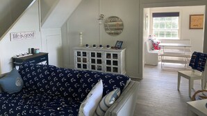 Un salón acogedor con un sofá azul, un banco blanco y una mesa de comedor. Se ve una escalera que sube a otro nivel, y un letrero en la pared que dice "life is good".