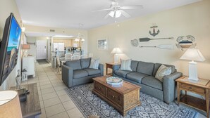 A compact living space with a sofa, coffee table, and a TV on a stand. A ceiling fan is visible, and there are nautical-themed wall decorations. - Ariel Dunes II 708 *Stunning Beach Views*Pools* (Miramar Beach)