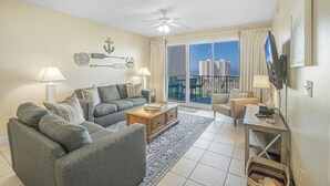 A living room with a gray sofa set, a wooden coffee table, and a TV on a stand. There is a large window with a view of buildings and a balcony. - Ariel Dunes II 708 *Stunning Beach Views*Pools* (Miramar Beach)