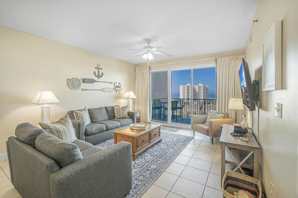 A living room with a gray sofa set, a wooden coffee table, and a TV on a stand. There is a large window with a view of buildings and a balcony.