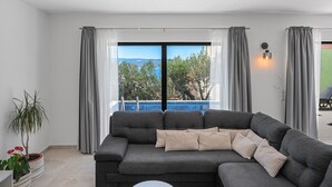 Un salon moderne avec un canapé d’angle gris, une petite table basse blanche et une vue sur une piscine à travers de grandes fenêtres.