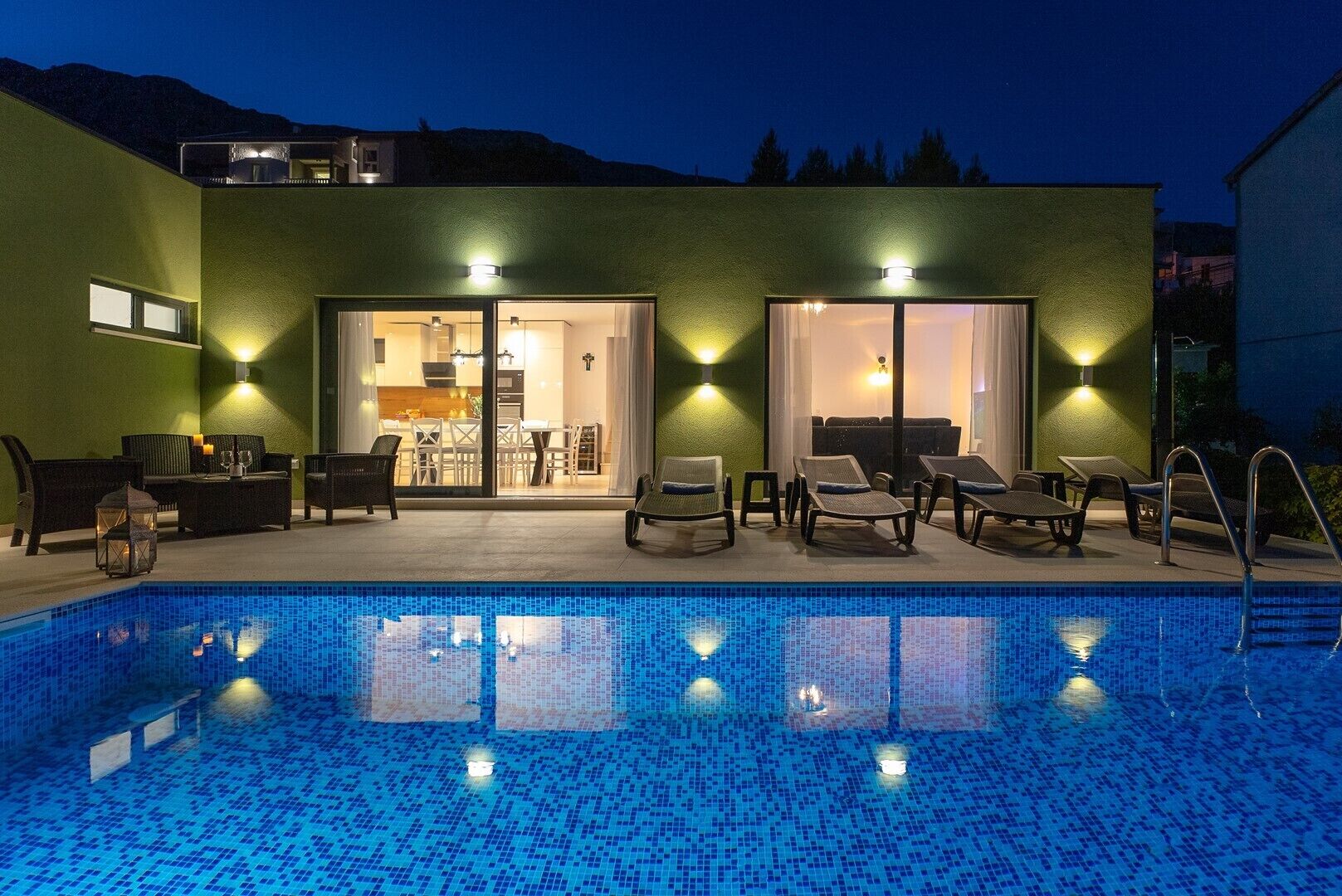 Área da piscina com espreguiçadeiras, uma piscina pequena e um edifício com fachada verde e portas de vidro que dão para o interior.