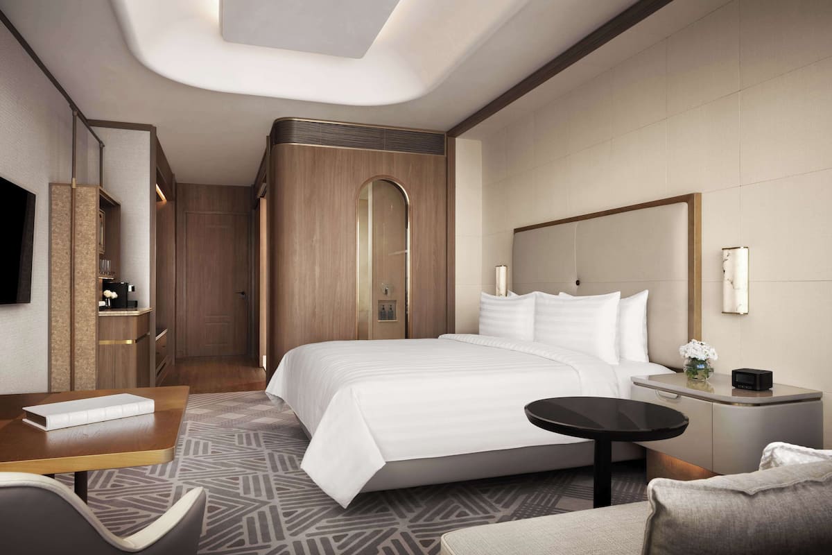 A modern hotel room with a large bed, a desk, a chair, a small table, a nightstand, and a TV.
