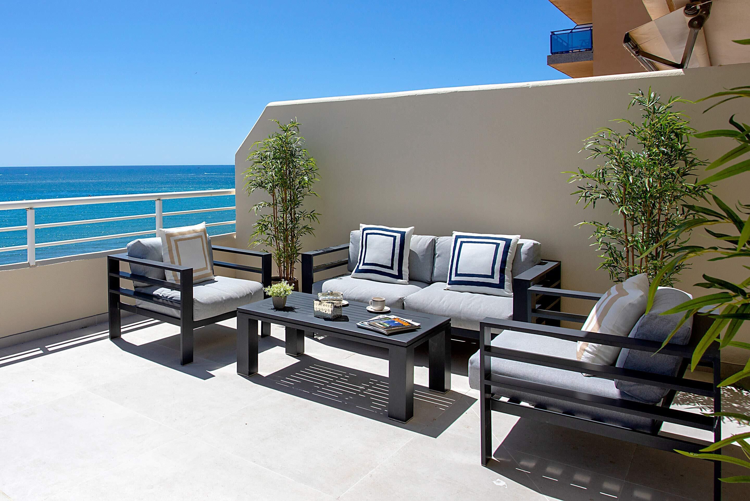Un balcon aménagé avec un canapé, des fauteuils et une table basse. On y trouve deux plantes en pot et une petite table sur laquelle sont posés un livre et une tasse. Le balcon offre une vue sur l’océan.