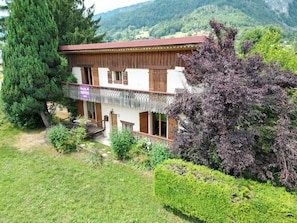 A traditional chalet with a wooden facade, a balcony with a wrought iron railing, and a view of the surrounding forest and mountains. - 🏡☀️ Chez Lily – Family House at the Foot of Le Criou 🌱⛰ (Samoëns)