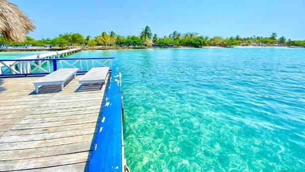 A wooden deck with lounge chairs extending over clear turquoise water, a thatched roof structure, and a distant shoreline with greenery and buildings. - Hotel Tintipan (Isla Tintipán)