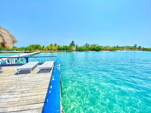 A wooden deck with lounge chairs extending over clear turquoise water, a thatched roof structure, and a distant shoreline with greenery and buildings. - Hotel Tintipan (Isla Tintipán)