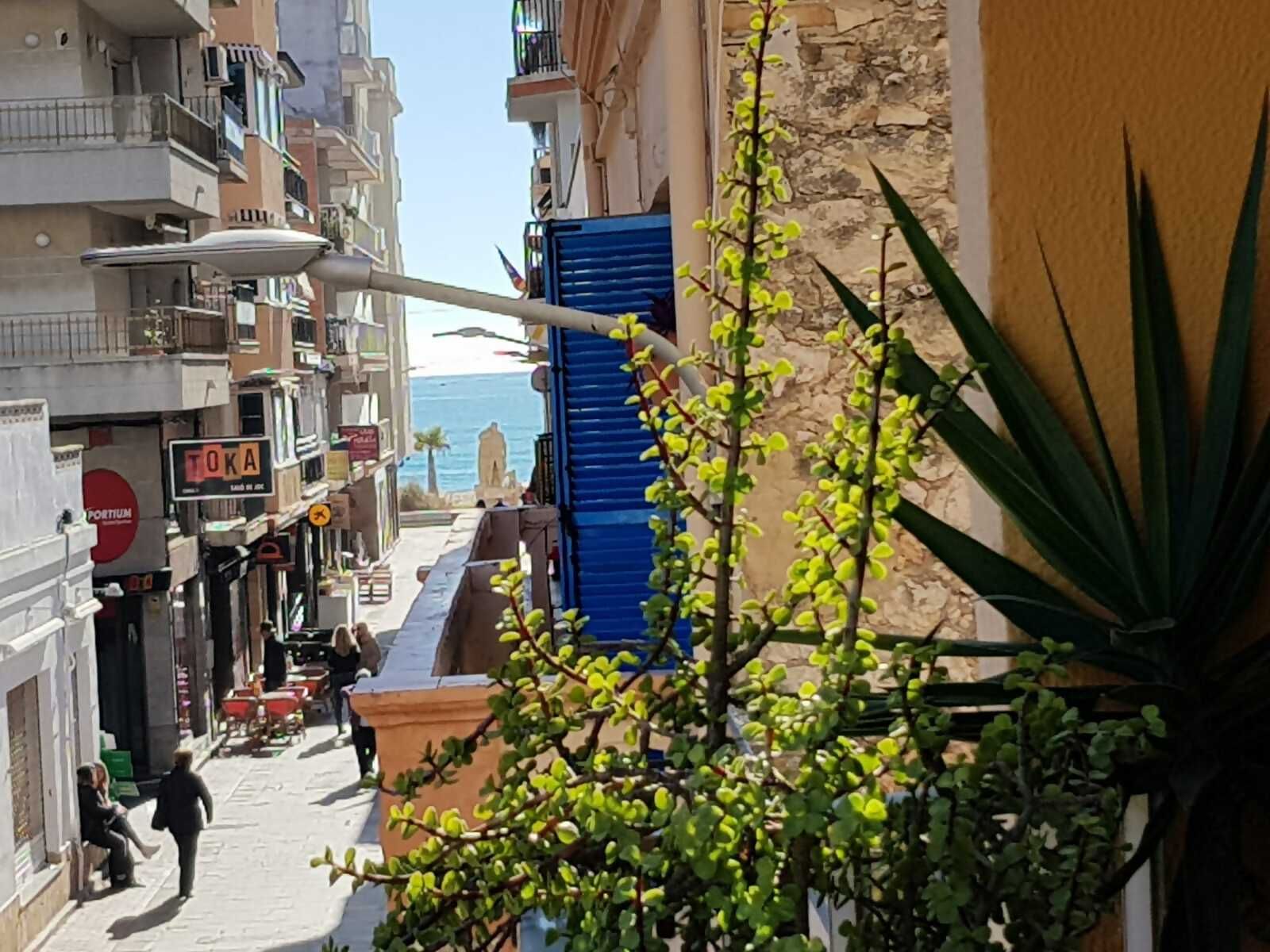 En smal gade med bygninger på begge sider, en butik med blå skodder, og en statue i det fjerne.