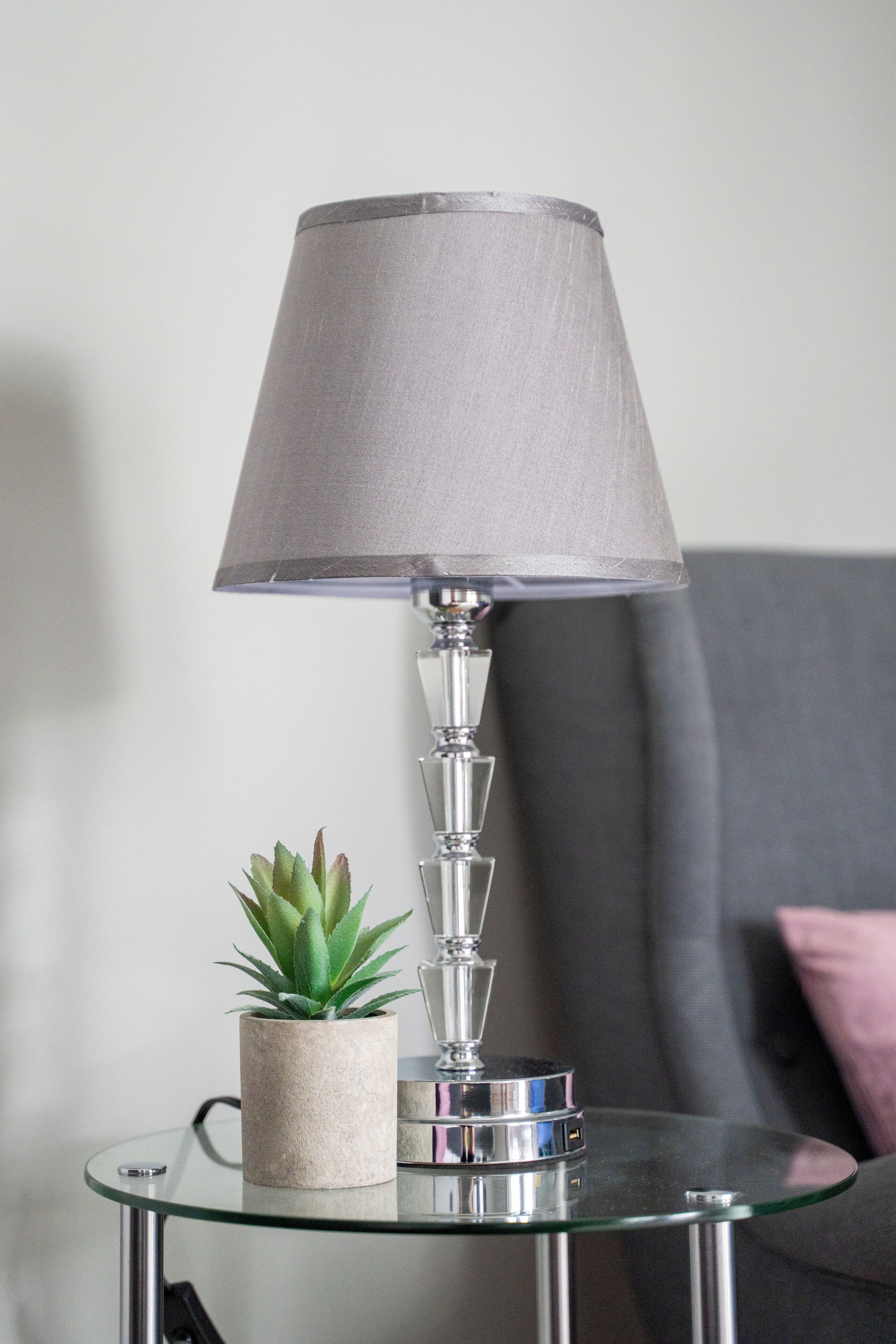 Een moderne lamp met een cilindrische voet en een conische kap, geplaatst op een glazen tafel met een kleine potplant ernaast.