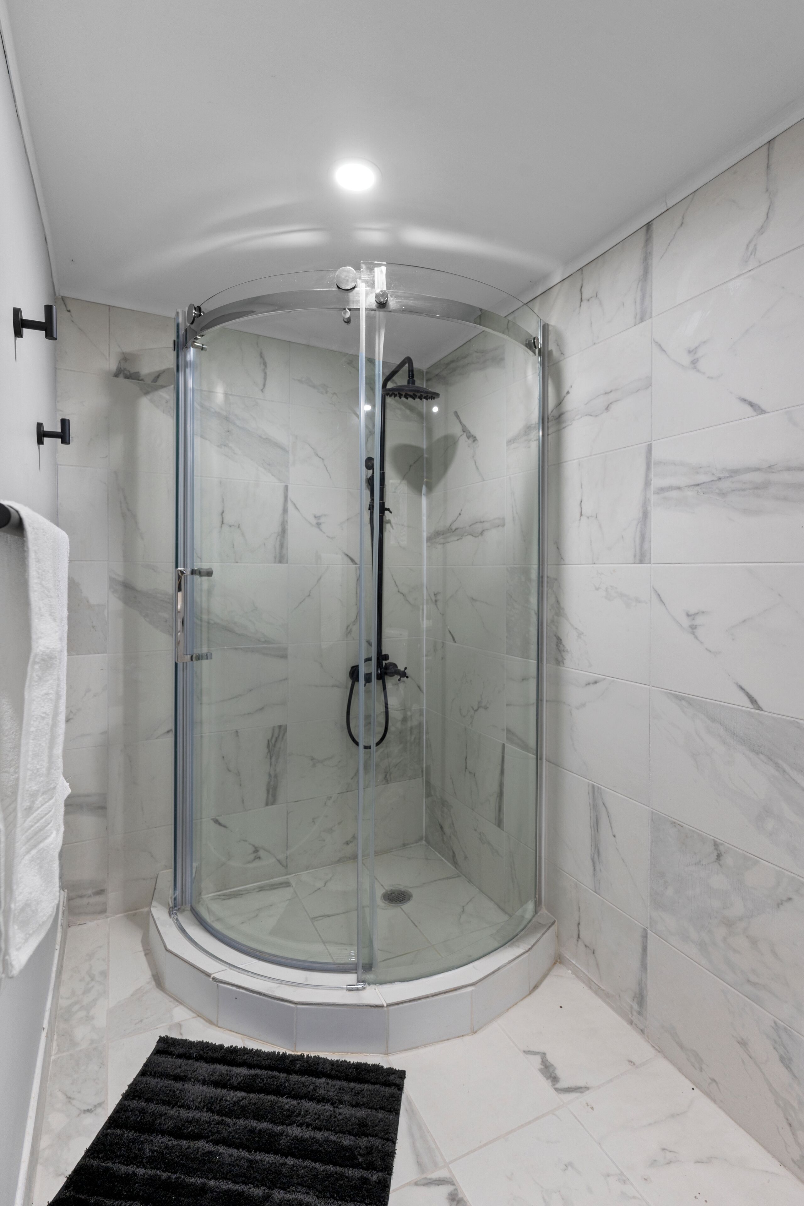 A modern bathroom with a glass-enclosed shower, white marble tiles, and a black rug.