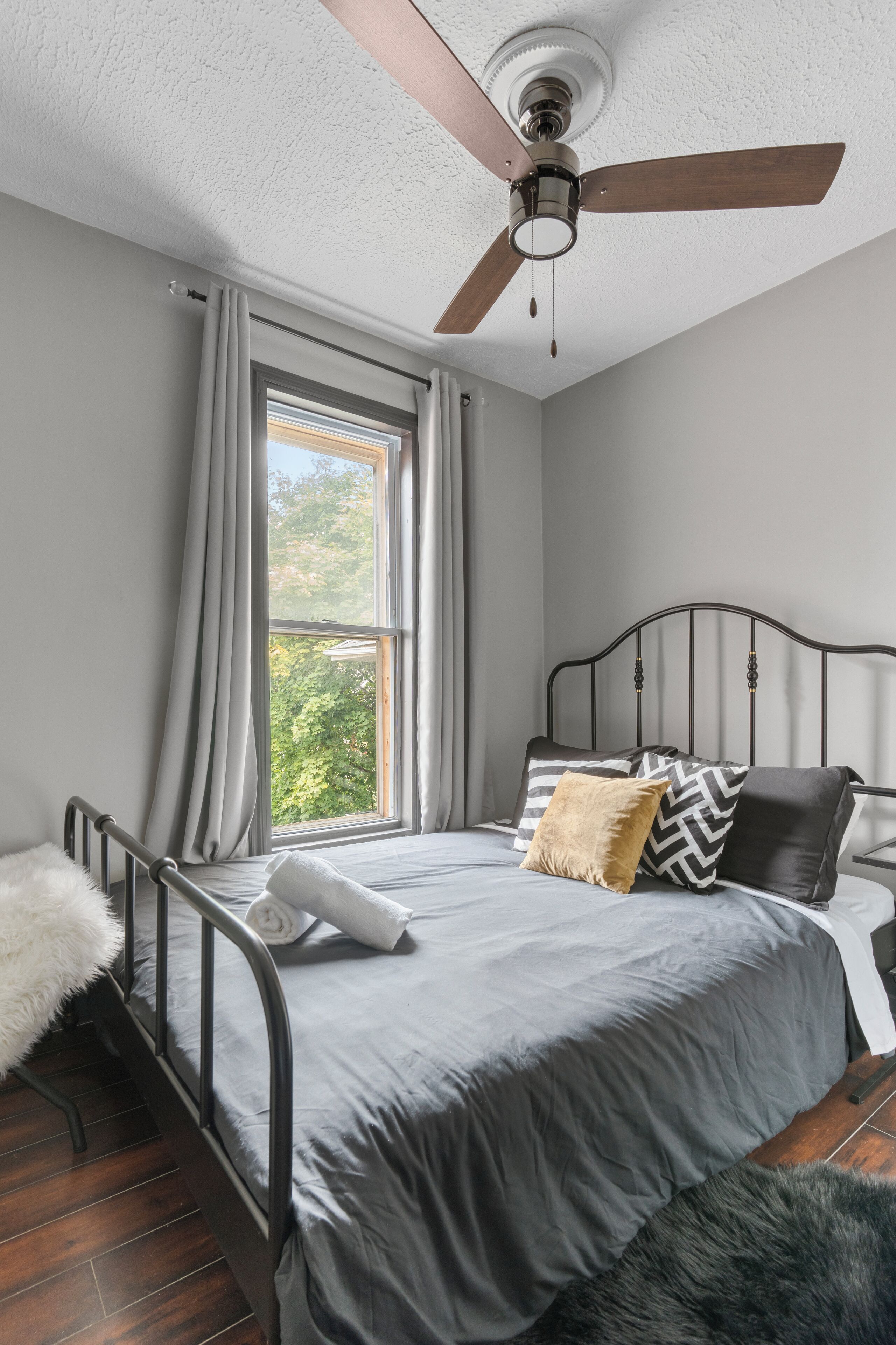 Ein Schlafzimmer mit Bett, Deckenventilator, Fenster mit Vorhängen und Blick auf Grünfläche.