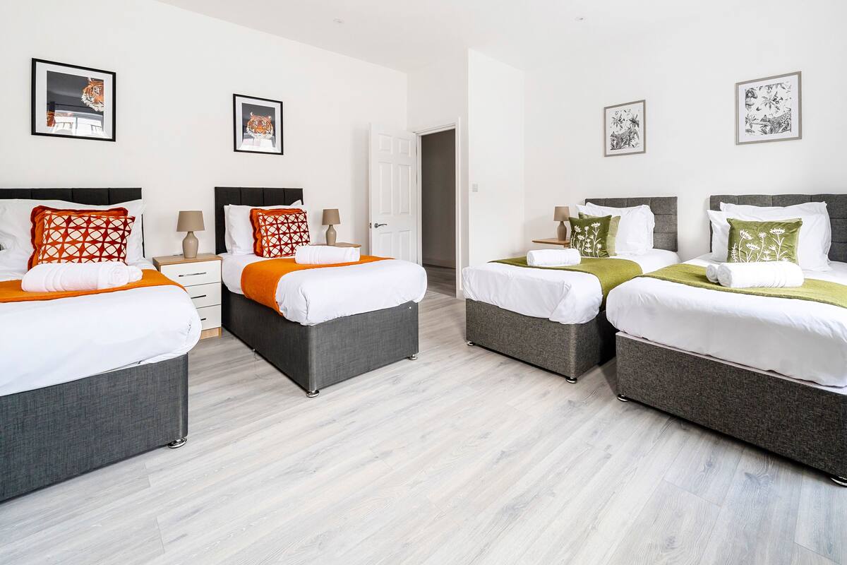 Ein Hotelzimmer mit zwei Betten, jeweils mit grauem Kopfteil, weißer Bettwäsche und Zierkissen. An der Wand hängt Kunstwerk, und eine Tür führt in einen weiteren Raum.