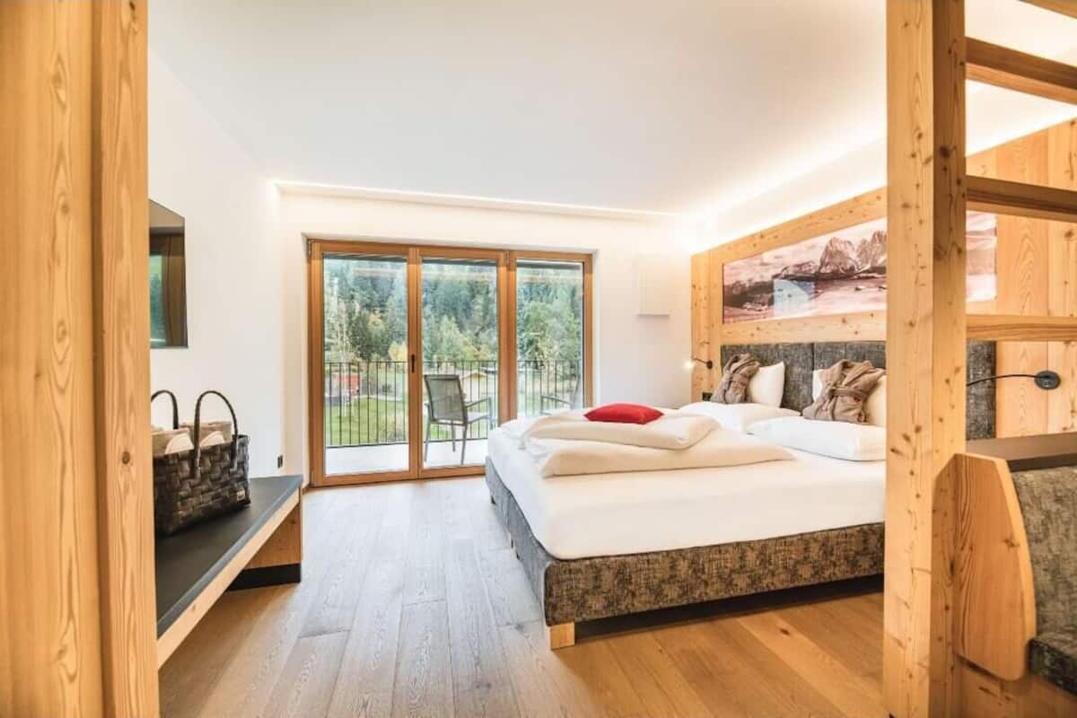 A bedroom with a large bed, a wooden headboard, a sliding glass door leading to a balcony, and a view of greenery.