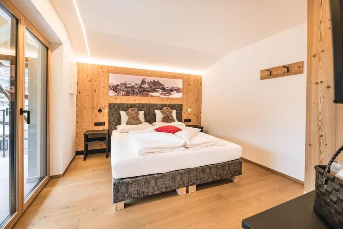 A modern bedroom with a wooden headboard, a large bed with white bedding, a red pillow, and a view of a snowy mountain through a window.