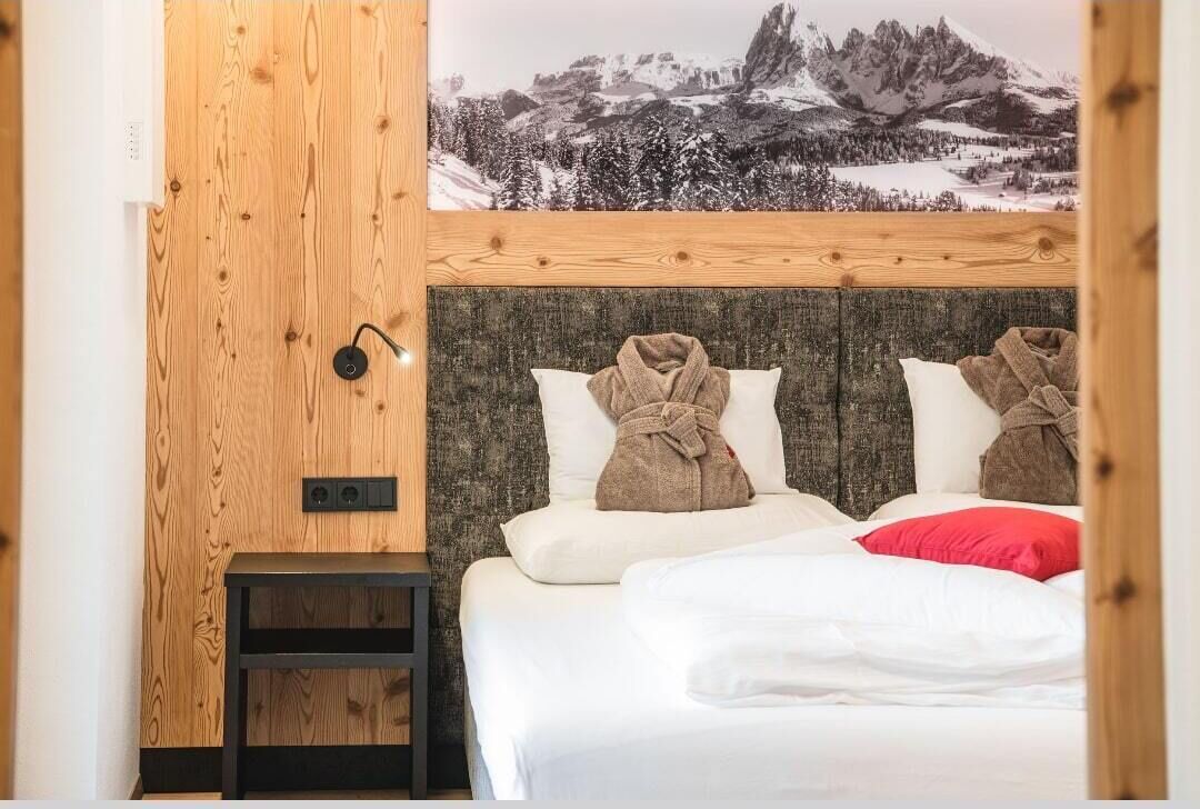 A hotel room with a wooden headboard, a bed with white bedding and two towels, a small bedside table, and a wall-mounted light.