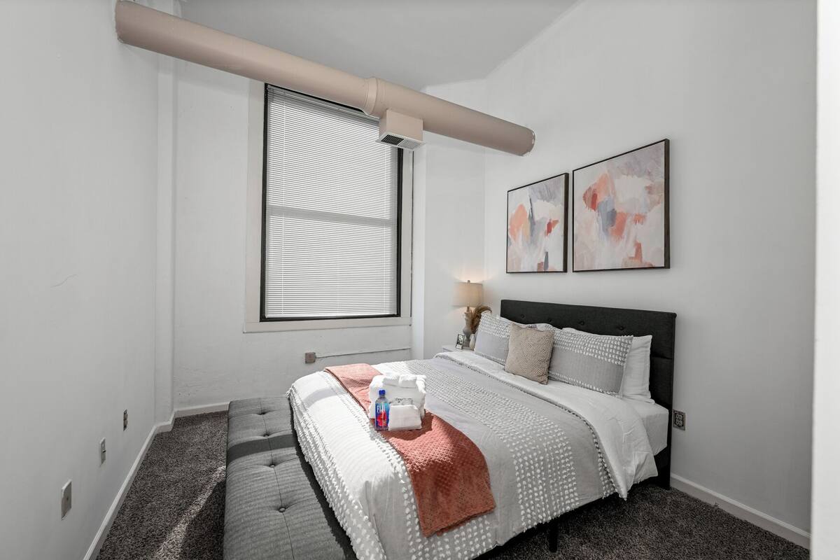 A bedroom with a bed, a window with blinds, a nightstand with a lamp, and abstract wall art.