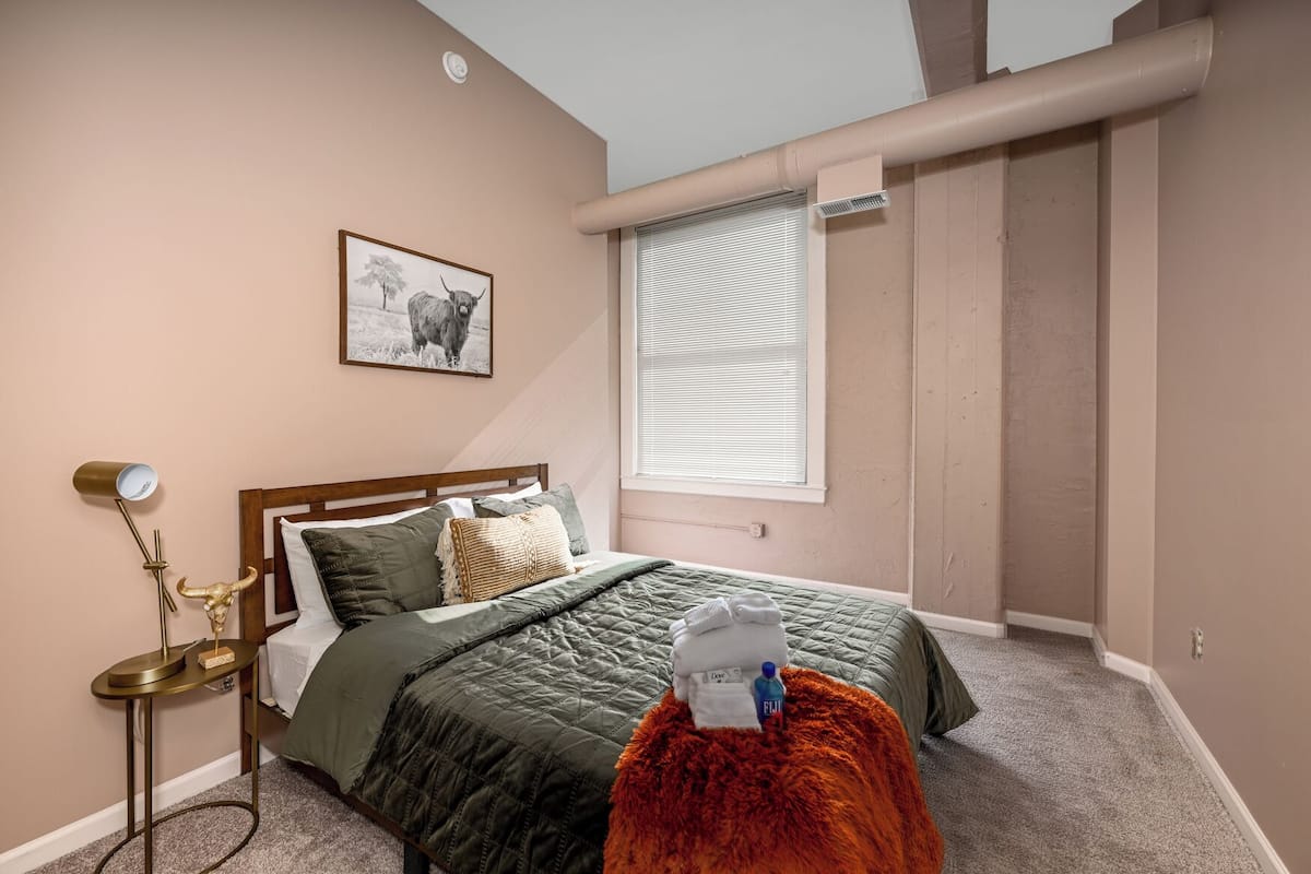 A bedroom with a bed, a bedside lamp, a framed picture on the wall, and a window with blinds.