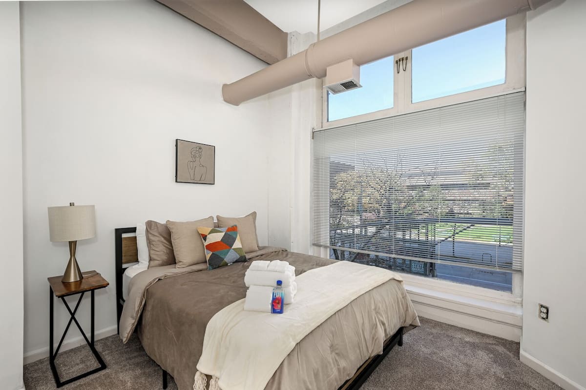 A modern hotel room with a large window, a bed with a beige blanket, a bedside lamp, and a colorful pillow.