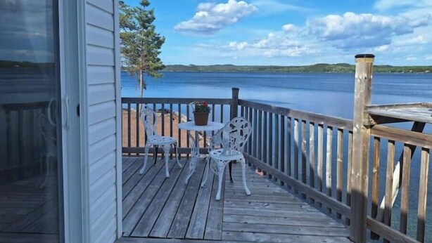 A wooden deck with a table and chairs, overlooking a calm body of water and distant land. - Skipper's View Cottage by the Sea - Pet Friendly! (Point Leamington)
