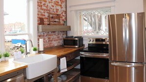 Een keuken met een bakstenen muur, roestvrijstalen apparatuur, een houten aanrechtblad en een raam met uitzicht op gebouwen.