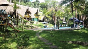 Um resort tropical com área de piscina, construções com telhados de palha, palmeiras e um parquinho.
