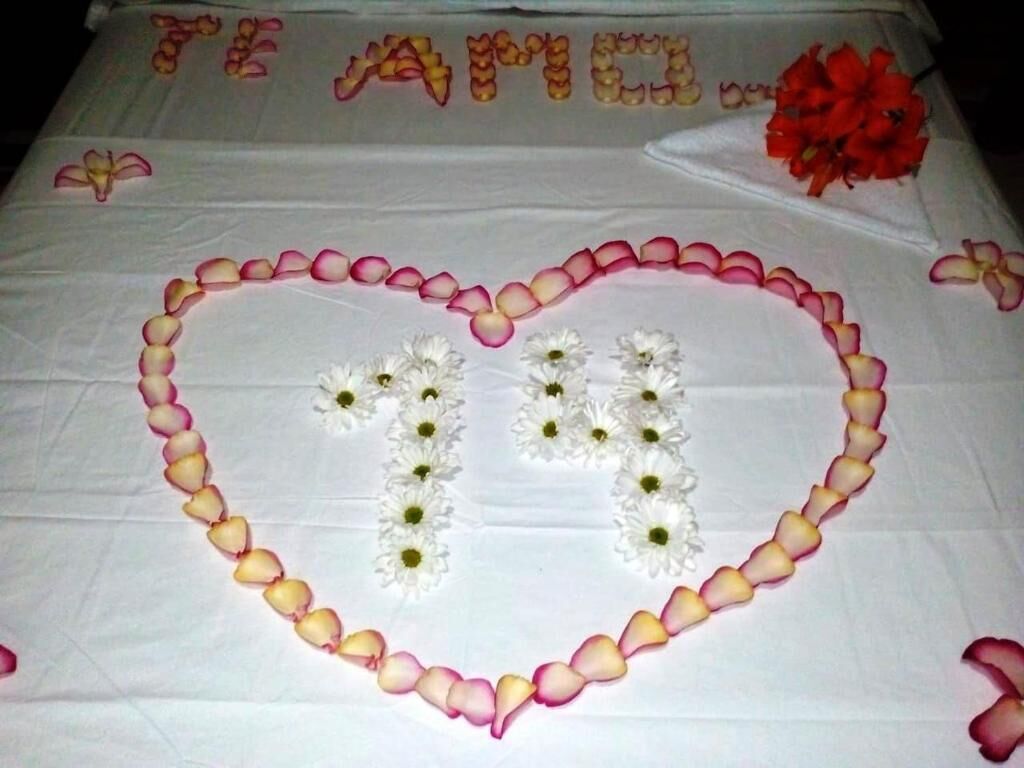 A bed with a heart-shaped arrangement of rose petals and white flowers, a red flower bouquet, and the text "TE AMO" on the bedspread.