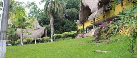 A tropical resort with thatched-roof buildings, lush greenery, and a well-maintained lawn.