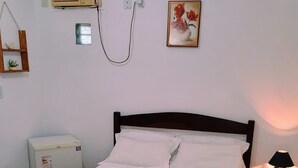 Una cama bien hecha con sábanas blancas y una manta marrón, una mesita de noche con una lámpara, un aire acondicionado de pared y un cuadro con flores enmarco.