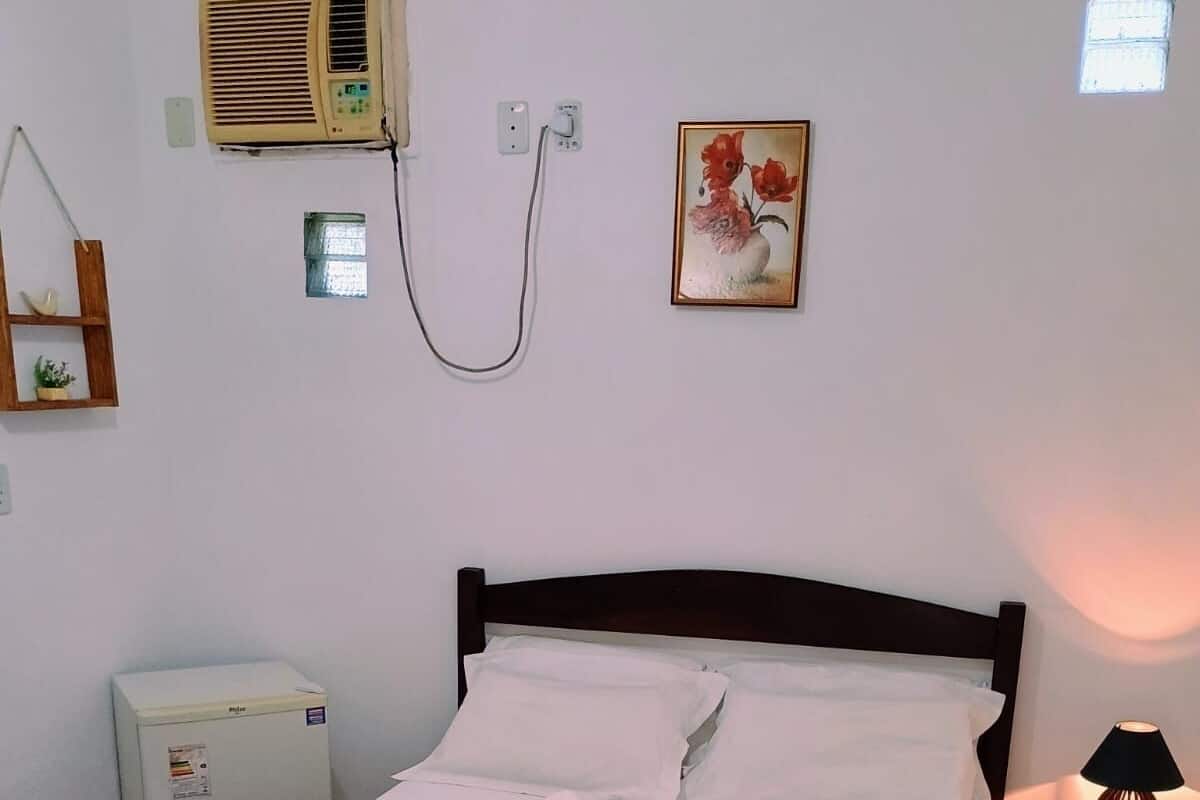 A neatly made bed with white linens and a brown throw blanket, a small bedside table with a lamp, a wall-mounted air conditioner, and a framed floral painting.