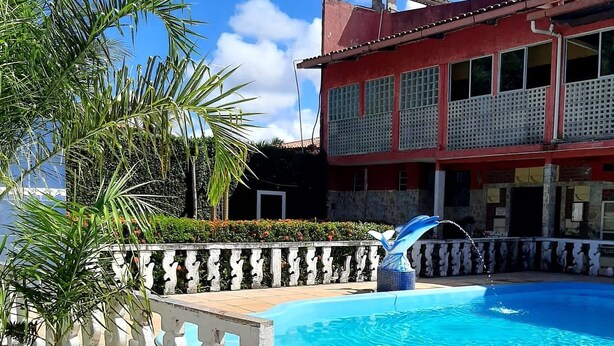 Una piscina con cerramiento blanco decorativo, palmeras y un edificio rojo con varias ventanas al fondo.