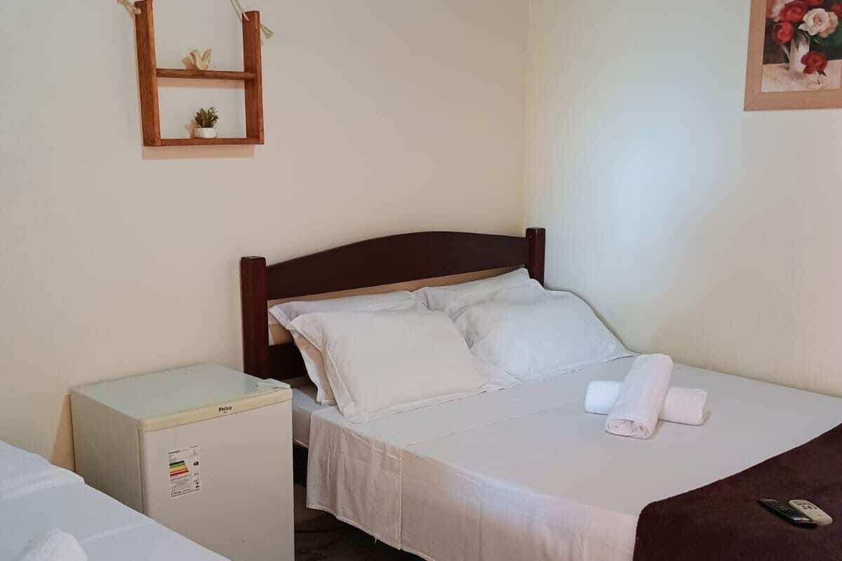 A hotel room with a bed, a small desk, a refrigerator, a wall-mounted shelf with a plant, and an air conditioning unit.