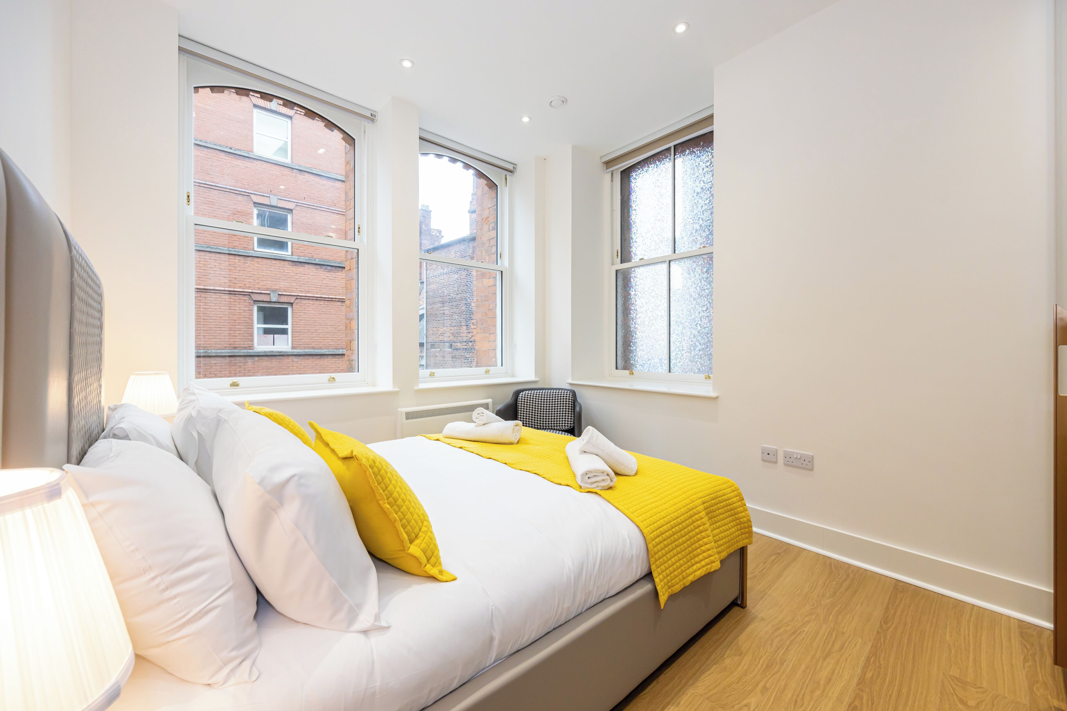 Un dormitorio moderno con una cama, dos ventanas, un radiador y una lámpara.