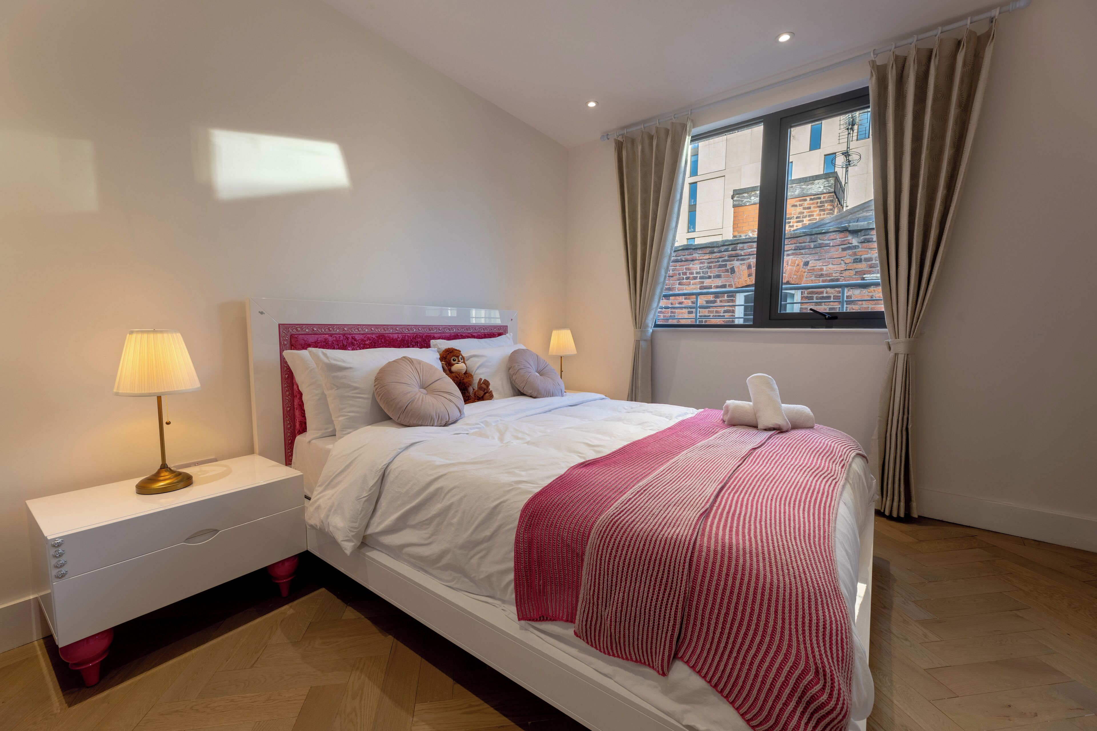 A neatly arranged bedroom with a bed, two bedside tables, a window with curtains, and a view of buildings outside.