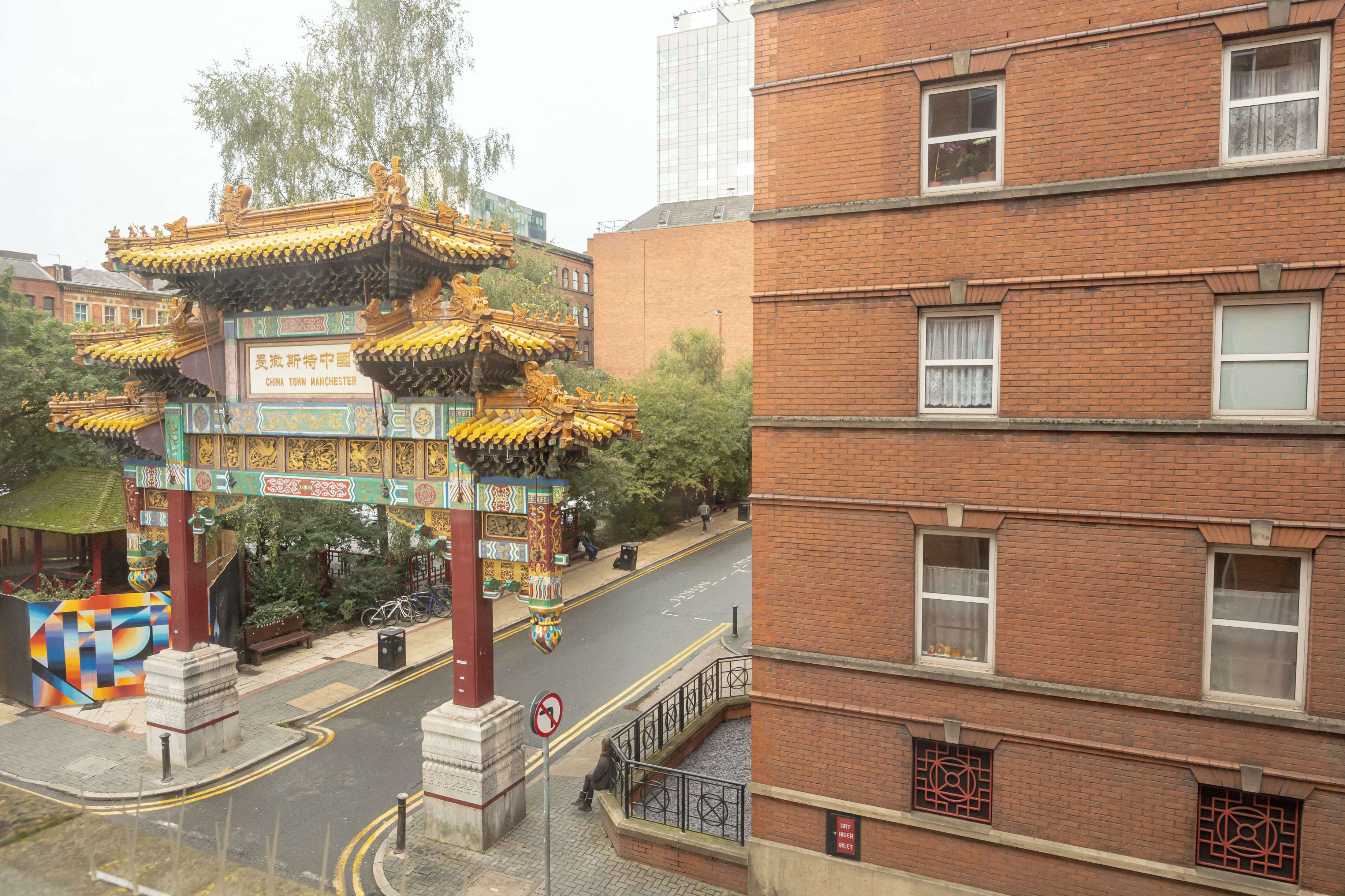 A traditional Chinese architectural structure with ornate details, a brick building with multiple windows, and a street with a no left turn sign.