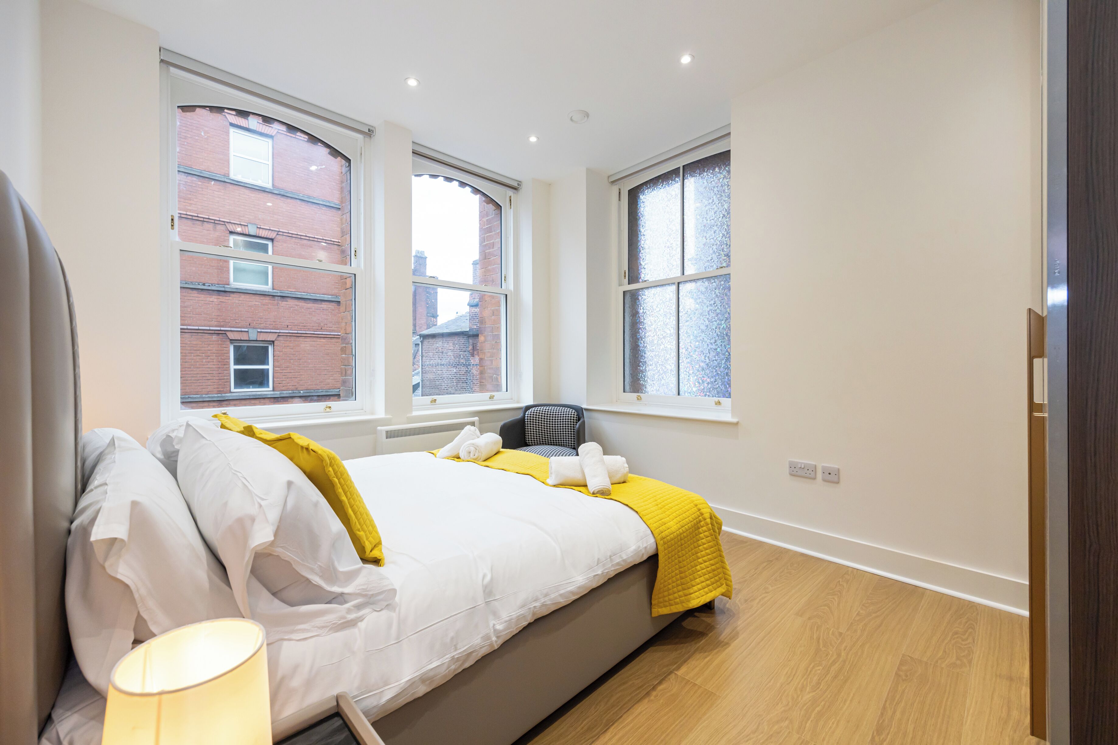 A modern bedroom with a bed, two windows, a chair, and a lamp.
