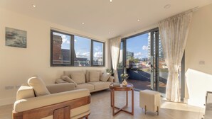 A modern living room with a sofa, a round wooden side table, and a view of the city through large windows. - Orient Residence Manchester (Manchester)