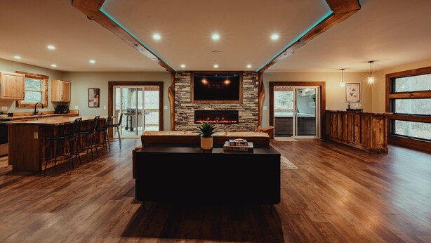 A spacious living area with a stone fireplace, wooden flooring, and a large window overlooking a deck.
