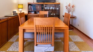 Un coin repas avec une table et des chaises en bois, une bibliothèque remplie de livres et d’objets décoratifs, un téléviseur fixé au mur et une petite table d’appoint avec une lampe.