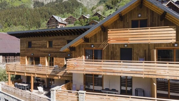 A wooden chalet with a balcony, multiple windows, and a stone foundation.