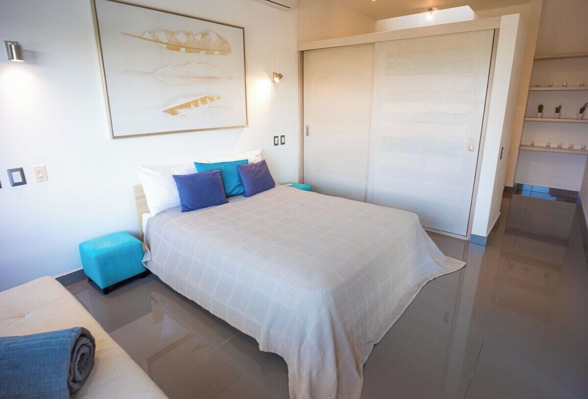 A modern bedroom with a large bed, a small side table, a blue ottoman, a wardrobe, and a framed artwork on the wall.