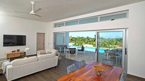 A modern living room with a sofa, a wooden dining table, and a television. There is a sliding glass door leading to an outdoor area with a pool.