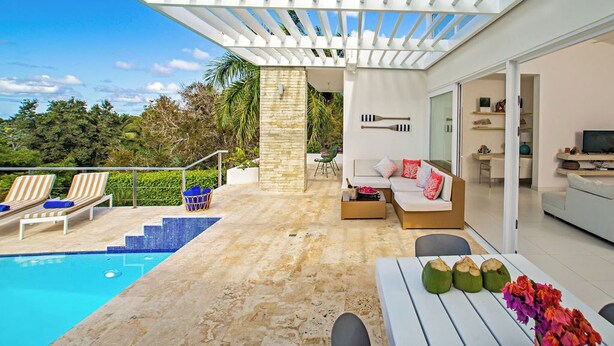 Un espacio exterior moderno con piscina, sillas de descanso, zona de estar con sofá y una pérgola que ofrece sombra.