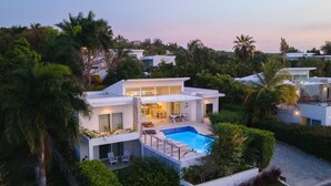 Una casa moderna de dos pisos con piscina, rodeada de abundante vegetación y palmeras.