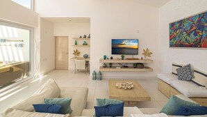 Una sala de estar moderna con un sofá, una mesa de centro y un televisor sobre un mueble. Hay objetos decorativos en estantes y un cuadro en la pared.