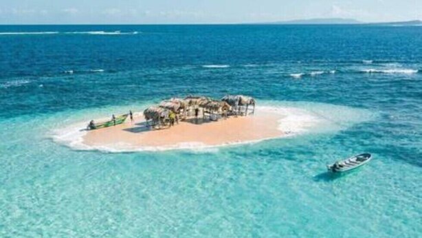 Eine Vogelperspektive zeigt eine kleine Insel mit Strand und einem strohgedeckten Gebäude, umgeben von klarem, türkisblauem Wasser.